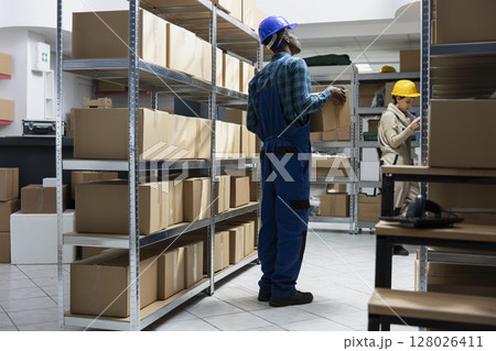 Black male employee examine labeled parcels on shelves in a storage unit depot. Logistics services support bulk shipping and retail inventory movement for supply chain, storehouse. 128026411