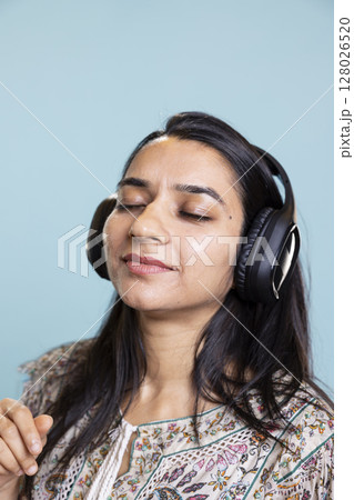 Happy woman playing electronic music and waving hand in the air, doing groovy dance moves in the studio. Cheerful confident girl wearing headphones and listening to her songs on camera. Happy woman playing electronic music and waving hand in the air, doing groovy dance moves in the studio. Cheerful confident girl wearing headphones and listening to her songs on camera. 128026520