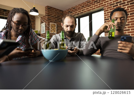 Male and female individuals focus on cellphones, browsing social media while sitting together at table with drinks and snacks. Diverse group of friends hanging out, engaged with mobile devices. 128026615