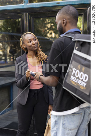 Black woman paying for her takeaway meal order with a contactless card at the doorstep. Delivery man carrying a thermal bag, completes fast food transaction using a wireless POS terminal. Black woman paying for her takeaway meal order with a contactless card at the doorstep. Delivery man carrying a thermal bag, completes fast food transaction using a wireless POS terminal. 128026700