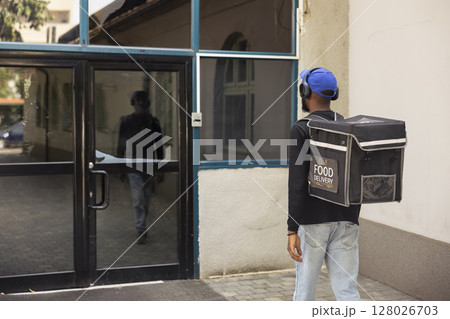Male courier walks down to reach client for a quick service food drop off, delivering food in his thermal backpack. Young man uses online delivery app for an express city service. Male courier walks down to reach client for a quick service food drop off, delivering food in his thermal backpack. Young man uses online delivery app for an express city service. 128026703
