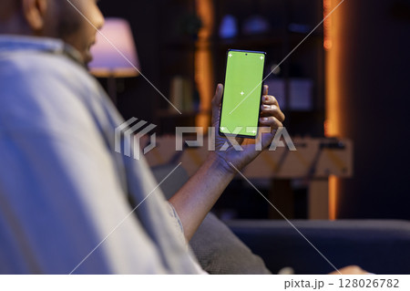 African american remote worker uses chroma key screen on gadget to work in the living room, sitting on the couch in his modern stylish living room. Happy young adult on the internet. African american remote worker uses chroma key screen on gadget to work in the living room, sitting on the couch in his modern stylish living room. Happy young adult on the internet. 128026782