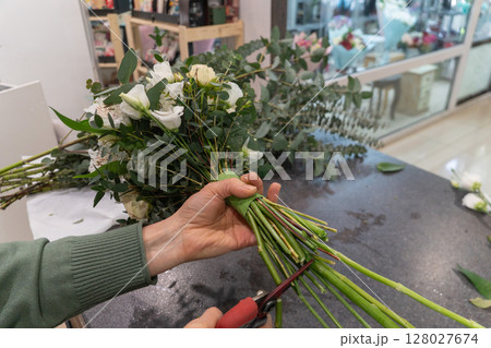 Close-up of florist trimming flower stems with pruning shears while holding a wrapped bouquet of white and peach flowers with eucalyptus greenery 128027674
