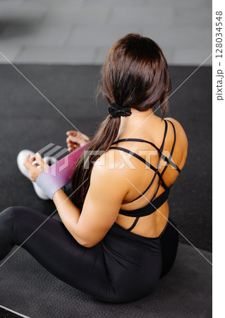 Young sporty woman wearing black sport suit performs exercises for the muscles of the hands, workout with resistance band on gym Young sporty woman wearing black sport suit performs exercises for the muscles of the hands, workout with resistance band on gym 128034548