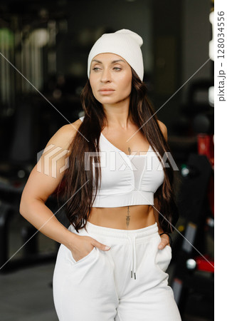 Beautiful fit woman looking at camera after gym exercise. Middle aged woman wearing sports bra and pants standing near simulator in gym Beautiful fit woman looking at camera after gym exercise. Middle aged woman wearing sports bra and pants standing near simulator in gym 128034556