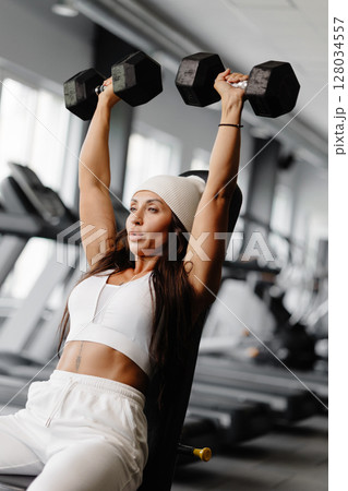 Athletic young fitness woman is wearing in white sport suit in gym outfit doing shoulder workout with dumbbells Athletic young fitness woman is wearing in white sport suit in gym outfit doing shoulder workout with dumbbells 128034557