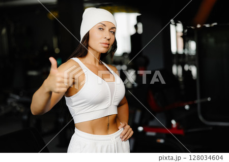 Portrait confident beautiful young brunette woman is wearing white suit and cap sitting on the gym 128034604