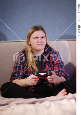 Teen girl sitting cross-legged on a bed, gripping a game controller with a focused and slightly frustrated expression during gameplay. 128037760