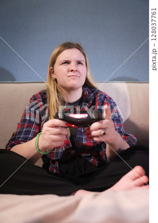 Focused teen girl plays video game at home, sitting cross-legged on a bed with a determined expression and holding a wireless controller. Focused teen girl plays video game at home, sitting cross-legged on a bed with a determined expression and holding a wireless controller. 128037761