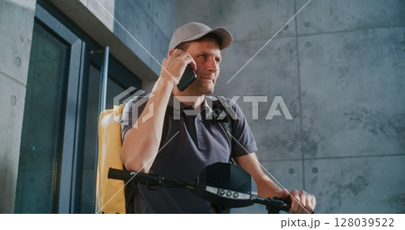Delivery Man on Electric Scooter Standing near Office, Accepting New Order from Client Talking by Phone Delivery Man on Electric Scooter Standing near Office, Accepting New Order from Client Talking by Phone 128039522