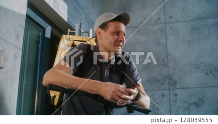 Tired Delivery Man Standing near Office, Searching for New Online Orders in App Using Smartphone Tired Delivery Man Standing near Office, Searching for New Online Orders in App Using Smartphone 128039555