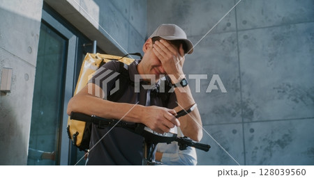 Tired Delivery Man Standing near Office, Searching for New Online Orders in App Using Smartphone Tired Delivery Man Standing near Office, Searching for New Online Orders in App Using Smartphone 128039560