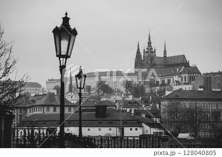 From a quiet street, the majestic Prague Castle looms in the background, framed by elegant lamp posts and the charm of historic rooftops. A captivating black and white view. 128040805