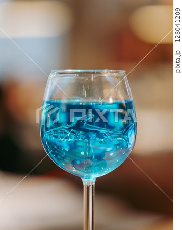 Glass of blue lagoon cocktail in shark high stem cocktail glass on beautiful bar background with bokeh. Aesthetic photo of blue lagoon cocktail in evening light, restaurant or bar interior 128041209
