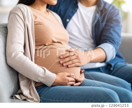 ソファに座る妊婦のお腹を優しく撫でる新婚夫婦の若いアジア人男性 ソファに座る妊婦のお腹を優しく撫でる新婚夫婦の若いアジア人男性 128041253