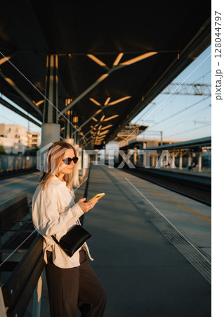 Young businesswoman waiting for train at sunset, using her smartphone on railroad station platform. Golden hour light casts shadows, enhancing urban travel experience. Businesswoman checking her phone 128044797