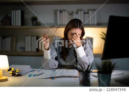 Late Night Stress. Young woman holding glasses while feeling overwhelmed at her desk. Late Night Stress. Young woman holding glasses while feeling overwhelmed at her desk. 128047349