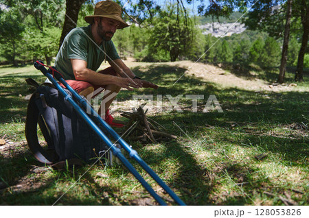 Man Preparing Bonfire in Forest Clearing with Backpack and Hiking Equipment Man Preparing Bonfire in Forest Clearing with Backpack and Hiking Equipment 128053826