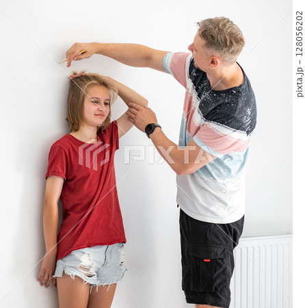 Happy Father Measures Smiling Daughter's Height And Marks It With A Pencil On The Wall Happy Father Measures Smiling Daughter's Height And Marks It With A Pencil On The Wall 128056202