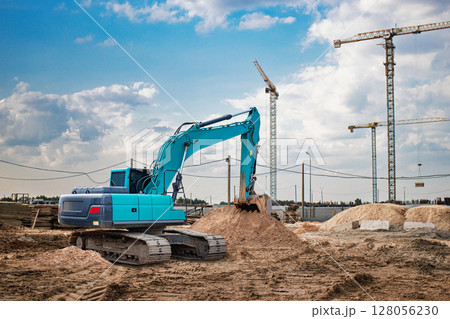 Excavator working at a construction site under a blue sky with cranes and scattered clouds Excavator working at a construction site under a blue sky with cranes and scattered clouds 128056230