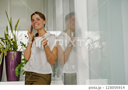 A cheerful woman talks on the phone while enjoying coffee in a cozy modern space 128056914