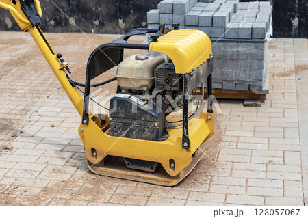 Vibratory rammer with vibrating plate on a construction site. Compaction of the soil before laying paving slabs. Close-up. Vibratory rammer with vibrating plate on a construction site. Compaction of the soil before laying paving slabs. Close-up. 128057067
