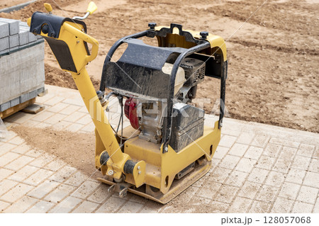 Vibratory rammer with vibrating plate on a construction site. Compaction of the soil before laying paving slabs. Close-up. Vibratory rammer with vibrating plate on a construction site. Compaction of the soil before laying paving slabs. Close-up. 128057068