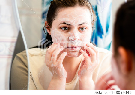 Woman removing makeup with face cleansing foam. Reflection in bathroom mirror. Beauty routine. Dermatology concept. Woman removing makeup with face cleansing foam. Reflection in bathroom mirror. Beauty routine. Dermatology concept. 128059349