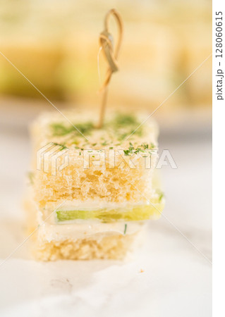 Soft white bread squares layered with a creamy cheese spread and crisp cucumber slices, topped with dried herbs and secured with bamboo cocktail picks. Perfect for elegant tea parties, bridal showers 128060615
