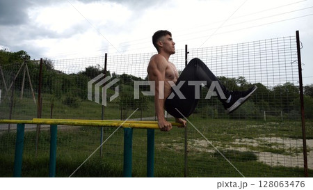 Strong guy training abdominal muscles on parallel bars at sports ground. Young shirtless sportsman exercising at nature. Sporty athlete working out at nature. Concept of active sportive lifestyle Strong guy training abdominal muscles on parallel bars at sports ground. Young shirtless sportsman exercising at nature. Sporty athlete working out at nature. Concept of active sportive lifestyle 128063476