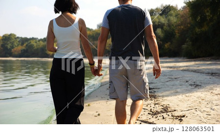 Romantic couple holding hands of each other and walking at coastline. Happy pair going along seashore at sunny day. Lovers spending time together at resort. Concept of vacation or holiday. Slow motion 128063501
