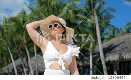 Young woman in white swimwear and straw hat walks along tropical beach 128064474