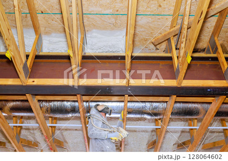 Worker in protective gear installs spray foam insulation with in exposed thermal barrier wooden framing at residential building. Worker in protective gear installs spray foam insulation with in exposed thermal barrier wooden framing at residential building. 128064602