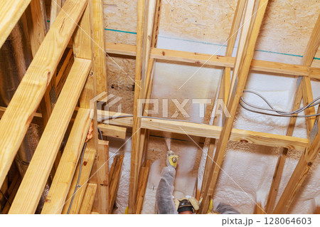 Skilled worker after install wooden beams, applies foam insulation material on ceiling framework in new house. Skilled worker after install wooden beams, applies foam insulation material on ceiling framework in new house. 128064603