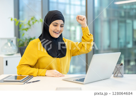 A woman in a hijab celebrates success in an office setting, smiling at her laptop. 128064684