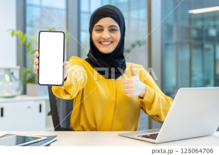 A smiling woman wearing a hijab shows a blank phone screen and gives a thumbs up, in an office setting. 128064706