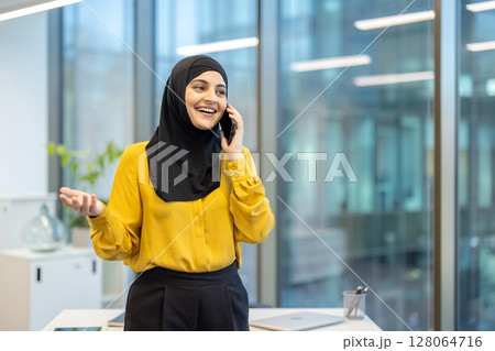 A cheerful woman wearing a hijab speaks on the phone in a bright office setting. 128064716