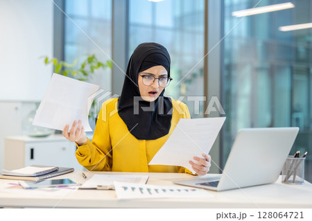 A shocked businesswoman in a hijab reviews paperwork in an office setting. The image portrays surprise and potential work-related stress. A shocked businesswoman in a hijab reviews paperwork in an office setting. The image portrays surprise and potential work-related stress. 128064721