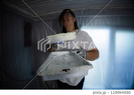 Woman painting a room with a roller brush, wearing protective gear. The space is covered in plastic sheeting, indicating a renovation or home improvement project in progress. 128064880