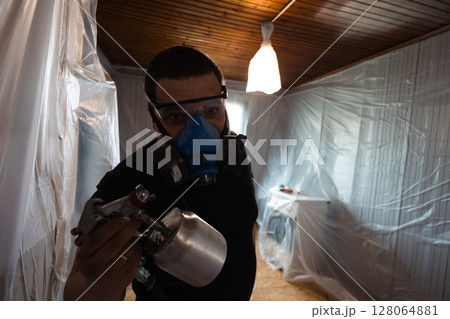 Painter wearing protective gear and using a spray gun for home renovation. The room is covered with plastic sheeting, indicating a focus on safety and cleanliness during the painting process. Painter wearing protective gear and using a spray gun for home renovation. The room is covered with plastic sheeting, indicating a focus on safety and cleanliness during the painting process. 128064881