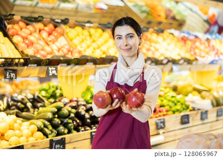 Young woman seller offering apples in vegetable shop Young woman seller offering apples in vegetable shop 128065967