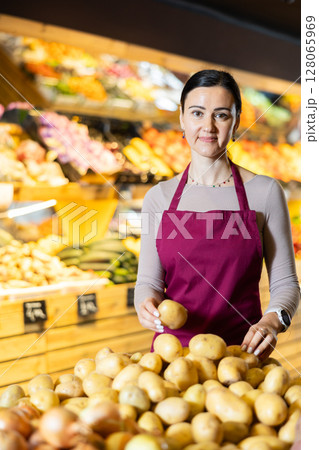 Middle-aged female seller putting potatoes on counter in grocery market Middle-aged female seller putting potatoes on counter in grocery market 128065969