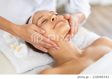 Young female client relaxing in spa salon during massage of face Young female client relaxing in spa salon during massage of face 128066341