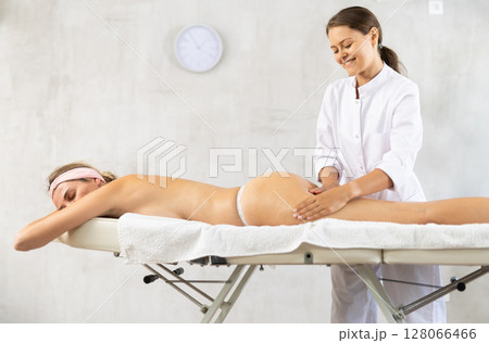 Smiling young professional masseuse performing thighs and buttocks massage to woman Smiling young professional masseuse performing thighs and buttocks massage to woman 128066466