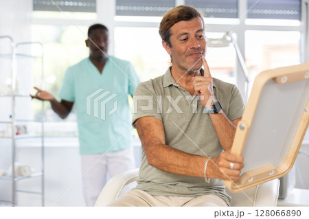 Man dissatisfied with results of rejuvenating at cosmetologist and meticulously examines reflection 128066890