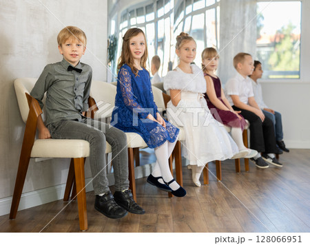 Little schoolchildren wearing elegant dresses sitting on chairs in school hall Little schoolchildren wearing elegant dresses sitting on chairs in school hall 128066951
