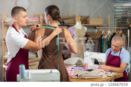 Guy tailor takes measurements from girl client, mature man work with sewing machine 128067039