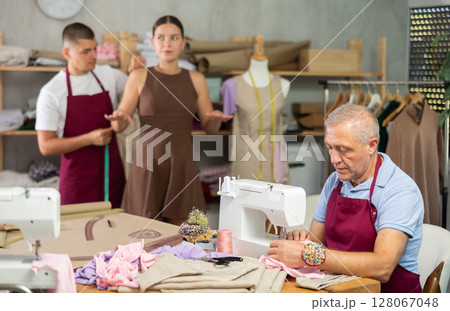 Tailor takes measurements from a woman next to an elderly employee of the atelier 128067048