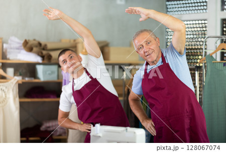 Elderly and young man doing warm-up exercises in sewing workshop Elderly and young man doing warm-up exercises in sewing workshop 128067074