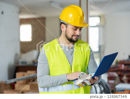 An architect in protective hard hat and yellow vest checks room renovation plan using laptop 128067349